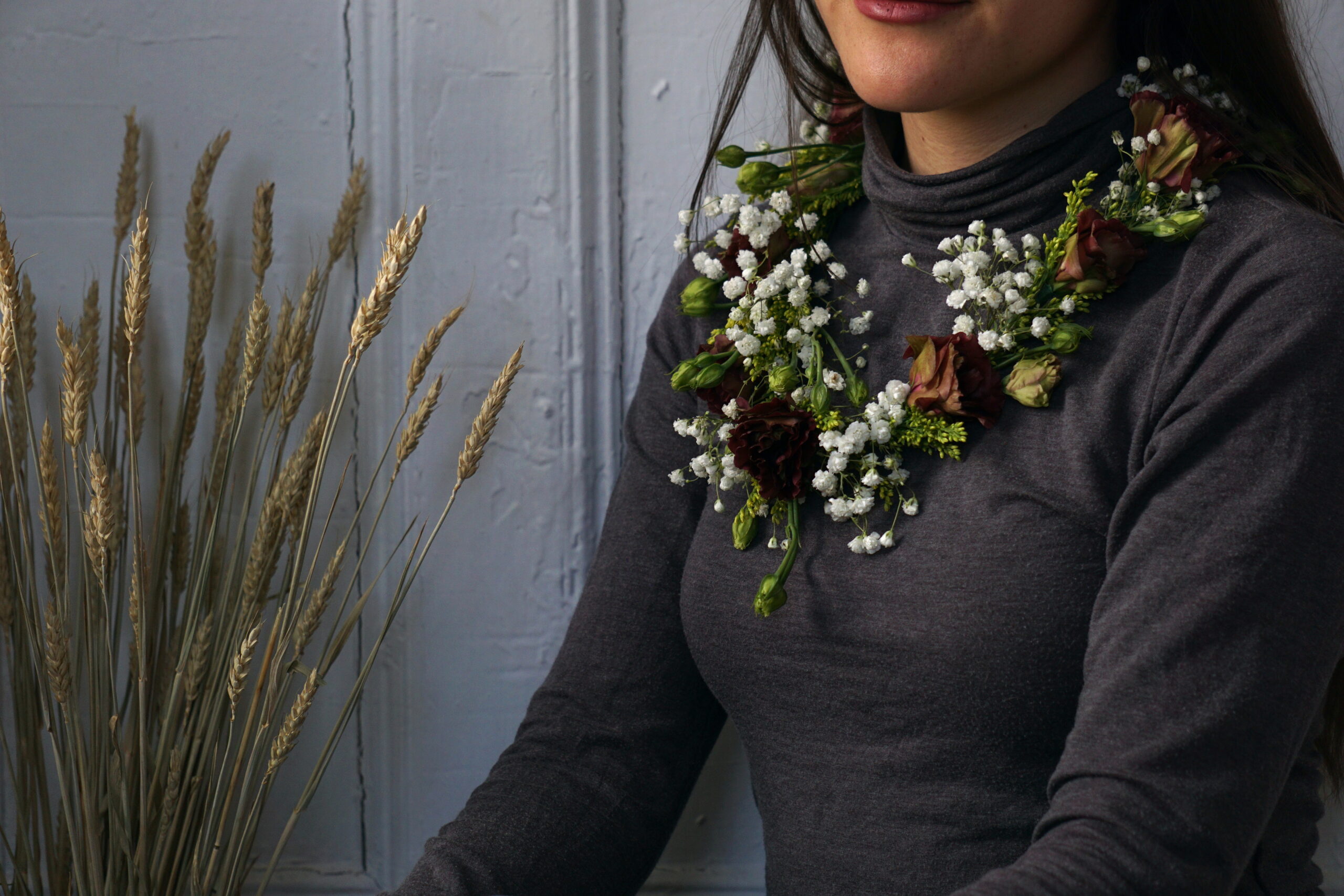 wheat grass arranged on left, woman with purple, green, and white floral wreath on her neck on right